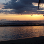 Waikiki at dusk, sailboats on the horizon beneath a cloud-streaked orange sky, the in-between hour between day and night