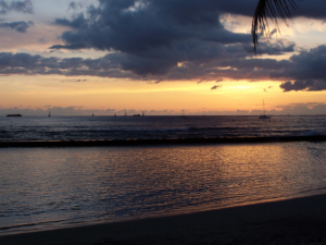 Waikiki at dusk, sailboats on the horizon beneath a cloud-streaked orange sky, the in-between hour between day and night